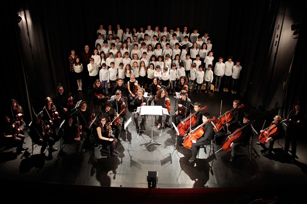 Coro numeroso de alunos no palco, acompanhado por uma orquestra